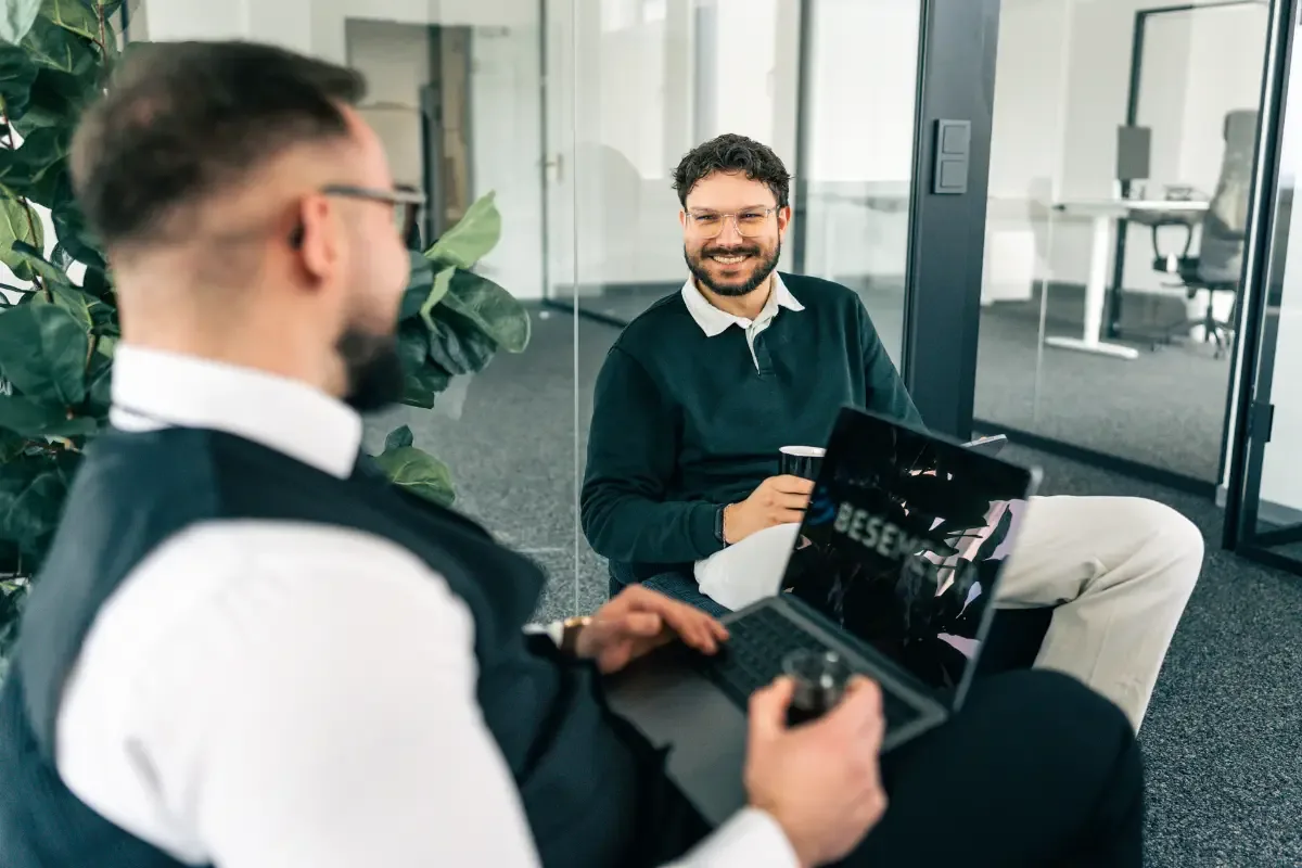 Zwei Mitarbeiter der Bese Media GmbH besprechen Zahlen am Laptop, einer sitzt am Tisch, der andere im Sessel